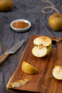 Durgun bir yaşam kompozisyonu, tahta bir kesme tahtasının üzerinde hafifçe tarçınla tozlanmış dilimlenmiş yeşil bir elmayı içerir. Bir bıçak ve küçük bir kase tarçın yumuşak odaklı arka planda gri bir bez üzerinde dinleniyor, birkaç tam elma ile birlikte.