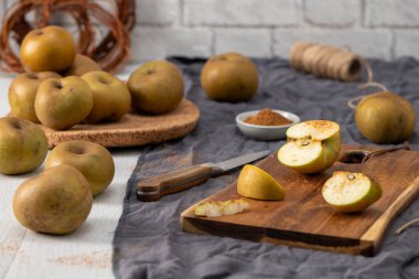 Kırsal bir natürmort, beyaz ahşap bir masada yeşil ve sarı benekli elma çeşitlerini yakalar. Bazıları mantar paspasın üzerindeyken, diğerleri ise bir bıçak ve küçük bir kase baharatla ahşap bir kesme tahtasının üzerinde tarçınla dilimlenmiş ve tozlanmış.