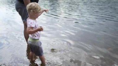 Küçük kız annesinin yanında duruyor ve suya taş atıyor.