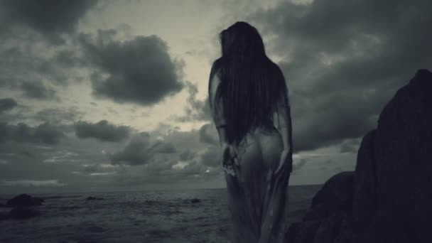 謎の女は美しい海と曇り夕焼け背景 黒と白のスローモーションのビデオを岩の近くの砂浜のビーチでドレスの夕日 バックの眺めの間にビーチで神秘的な美しさ — ストック動画