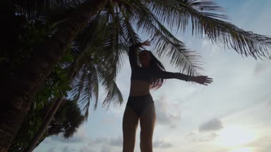 Güzel kadın kumsalda dans / alt görünümü içinde siyah mayo palmiye ağacı ve gökyüzü arka plan - ağır çekimde video üzerinden günbatımı sırasında kumsalda dans güzel kadın