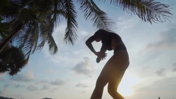 ビーチで踊る美人 ヤシの木と空背景 スローモーションのビデオの上に日没時に砂浜で踊る黒い水着で美しい女性観を底 — ストック動画