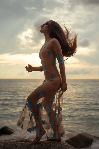 美丽的神秘女人穿着长裙在沙滩附近的岩石上海和多云的日落天空背景 — 图库照片