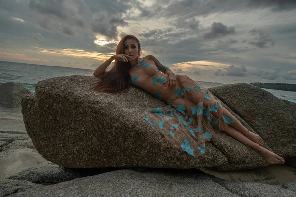 美丽的神秘女人穿着长裙躺在海中的岩石和多云的日落天空背景 — 图库照片