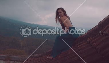 Oturan kırmızı ev çatı üzerinde / güzel güzel kadın oturan uzun mavi elbiseli kadın döşeli kırmızı inanılmaz dağ manzaralı ve bulutlu gökyüzü arka plan - ağır çekimde video karşı evin çatısı