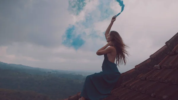 美丽的女人穿着长裙坐着蓝色的烟雾在瓷砖红色屋顶的房子反对惊人的山景和多云的天空背景 — 图库照片