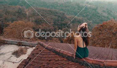 Kırmızı ev çatı katında oturan güzel kadın / hava dron görünümü oturan uzun mavi elbiseli güzel kadının döşeli kırmızı inanılmaz dağ manzaralı ve bulutlu gökyüzü arka plan - ağır çekimde video karşı evin çatısı