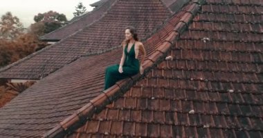 Kırmızı ev çatı katında oturan güzel kadın / hava dron görünümü oturan uzun mavi elbiseli güzel kadının döşeli kırmızı inanılmaz dağ manzaralı ve bulutlu gökyüzü arka plan - ağır çekimde video karşı evin çatısı