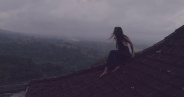 Kırmızı ev çatı katında oturan güzel kadın / hava dron görünümü oturan uzun mavi elbiseli güzel kadının döşeli kırmızı inanılmaz dağ manzaralı ve bulutlu gökyüzü arka plan - ağır çekimde video karşı evin çatısı