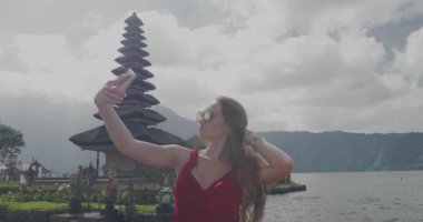 Genç kadın turist onu tatil Adası Bali, Endonezya - video yavaş hareket üzerinde Ulun Danu Batur Tapınağı'nda cep telefonu ile selfie fotoğraf yapıyor sahiptir