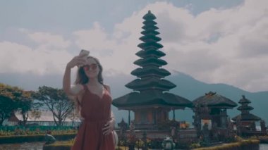 Genç kadın turist onu tatil Adası Bali, Endonezya - video yavaş hareket üzerinde Ulun Danu Batur Tapınağı'nda cep telefonu ile selfie fotoğraf yapıyor sahiptir
