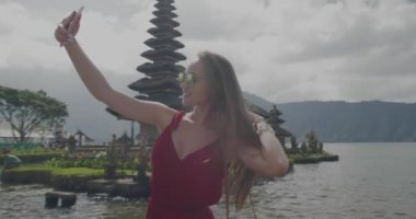 Genç kadın turist onu tatil Adası Bali, Endonezya - video yavaş hareket üzerinde Ulun Danu Batur Tapınağı'nda cep telefonu ile selfie fotoğraf yapıyor sahiptir