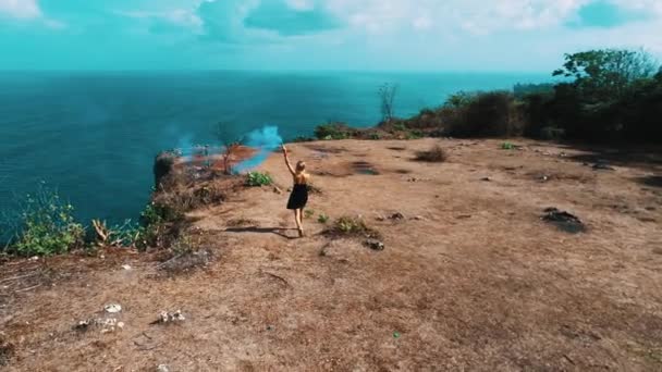 空中无人机拍摄漂亮的女孩玩彩色烟雾在悬崖上与惊人的海洋和多云的天空视图 视频慢动作 — 图库视频影像