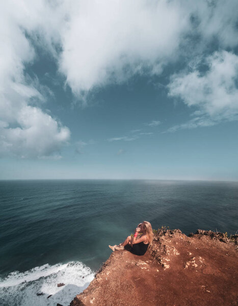 Pretty girl sitting on the cliff with amazing ocean and cloudy sky view 