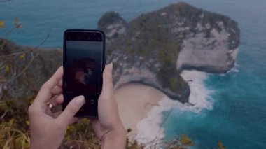 Kadın eller ile hareket eden telefon fotoğraf güzel Kelingking Beach kayalıklarla Nusa Penida, Bali, Endonezya yapıyor