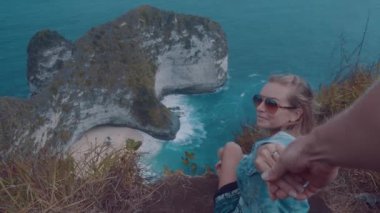 Cliff üzerinde oturan ve güzel Kelingking Beach kayalıklarla arka planda Nusa Penida, Bali, Endonezya üzerinden erkek arkadaşının elini tutarak kız