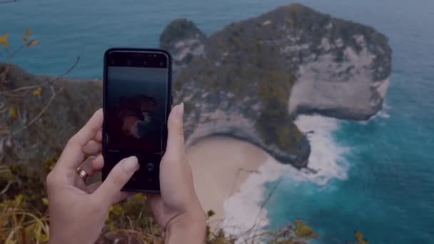 ペニダ島 バリ島 インドネシアの美しい崖 Kelingking ビーチの写真を行う携帯電話で女性の手をクローズ アップ — ストック動画