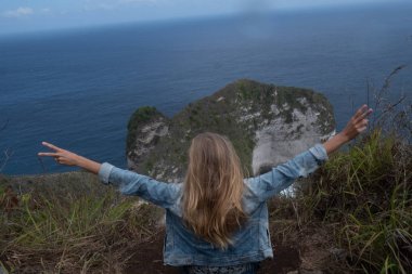 Cliff üzerinde oturan ve güzel Kelingking Beach kayalıklarla arka planda Nusa Penida, Bali, Endonezya üzerinde eller yükselterek arkadan görünüm
