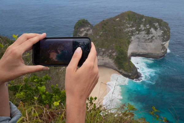 ペニダ島 インドネシア バリ島で崖を Kelingking ビーチの写真を行う携帯電話でクローズ アップ手 — ストック写真