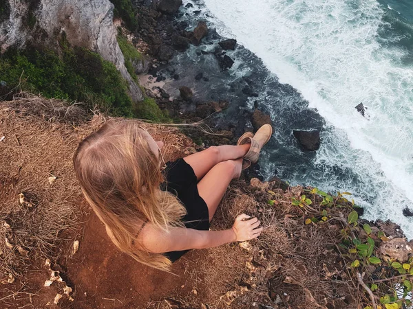 Top view of blonde woman tourist sitting on the cliff and admire beautiful ocean view