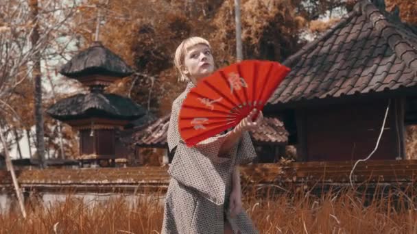 美しいファッションの少女赤い手ファンのポーズと着物の伝統的なバリの寺院の近くの屋外 屋外のスタイリッシュな流行に敏感な女性の肖像画 — ストック動画