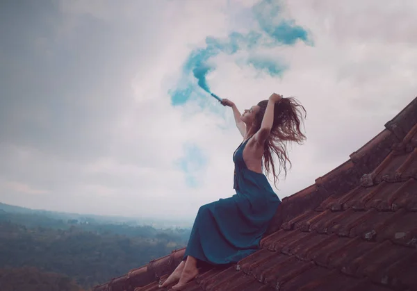 美丽的女人穿着长裙坐着蓝色的烟雾在瓷砖红色屋顶的房子反对惊人的山景和多云的天空背景 — 图库照片