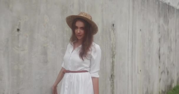 白いドレスと灰色のコンクリート壁で分離された麦わら帽子で微笑んでいる女の子のファッション美容肖像画 — ストック動画
