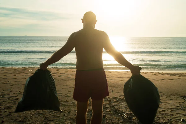 Back view of man tourist standing on the beach with collected bags of rubbish over sea and sunset background