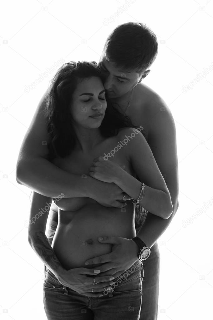 Intimate portrait of beautiful pregnant couple standing together over window background. Couple is topless and husband covers wife's breast with hand - black and white image