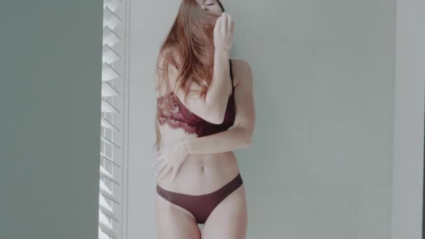 ランジェリー スローモーションのビデオ 晴れた日の午前中にポーズのブラインドと窓の近くに美しい赤い髪の少女 — ストック動画