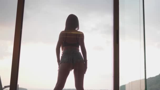 性感的黑发女人在牛仔短裤和胸罩跳舞在室外游泳池在多云的天空背景 从公寓内部透过露台窗口观看 慢动作视频 — 图库视频影像
