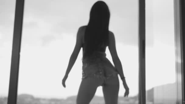 デニムのショート パンツとブラジャーをスローモーションで曇り空の背景 黒と白のビデオの上テラス窓の近くダンスでセクシーなブルネットの女性の背面図 — ストック動画