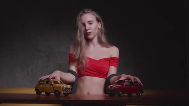 赤トップではかなり若いブロンドの女性は灰色の壁の背景の上テーブルにおもちゃの車で遊ぶ — ストック動画