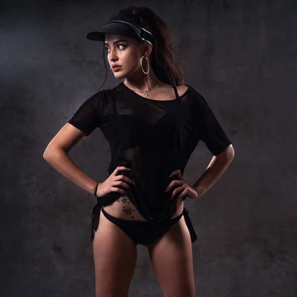 Sensual woman wearing black see through t-shirt, lingerie and sun visor posing in the studio with grey concrete walls