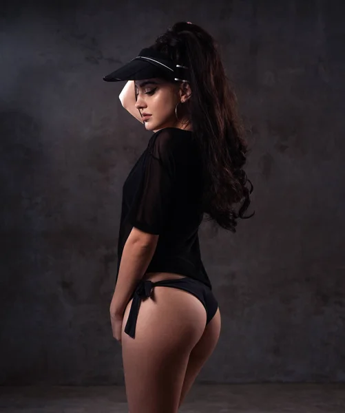 Sensual woman wearing black see through t-shirt, lingerie and sun visor posing in the studio with grey concrete walls