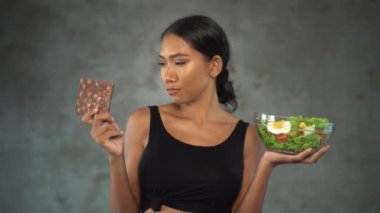 Beton arka plan üzerinde sağlıklı salata ile çikolata ve kase tutan siyah t-shirt güzel genç gülümseyen kadın Portresi. Sağlıklı ve sağlıksız gıda arasında seçim Pretty genç kadın