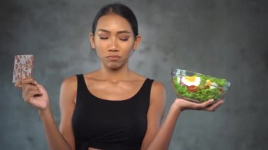 Beton arka plan üzerinde sağlıklı salata ile çikolata ve kase tutan siyah t-shirt güzel genç gülümseyen kadın Portresi. Sağlıklı ve sağlıksız gıda arasında seçim Pretty genç kadın