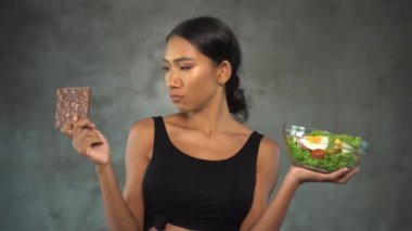 Beton arka plan üzerinde sağlıklı salata ile çikolata ve kase tutan siyah t-shirt güzel genç gülümseyen kadın Portresi. Sağlıklı ve sağlıksız gıda arasında seçim Pretty genç kadın