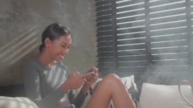 Gülümseyen güzel kız cep telefonu ile evde kanepede oturan. İnsanlar, rahat yaşam tarzı ve teknoloji konsepti