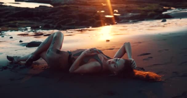 美丽的性感女人在闪闪发光的泳衣在黑色沙滩在惊人的金色日落中摆姿势 视频在慢动作 — 图库视频影像