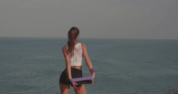 日の出時の海岸崖で抵抗力帯で運動する若い魅力的な女性のバックビュー — ストック動画