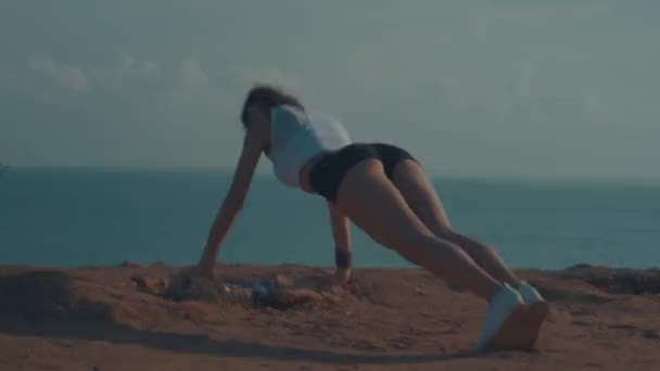 日の出時に海岸の崖で板の膝から胸の運動をしている若い魅力的な女性 — ストック動画