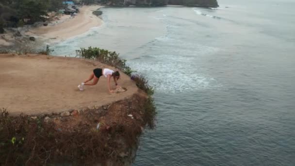 日の出時の海岸の崖で板膝から胸の運動をしている若い魅力的な女性の空中ドローンビュー — ストック動画