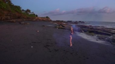 Gün batımında siyah kum plajda renkli duman ile bikini dans güzel şehvetli kadın havadan drone görünümü