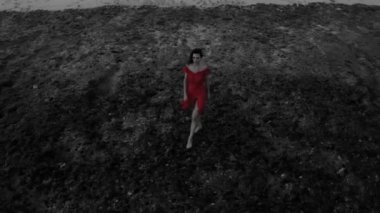 Gün batımında kumlu plaj yakınındaki kayalar üzerinde poz kırmızı elbiseli güzel kadının havadan drone görünümü - siyah, beyaz ve kırmızı video