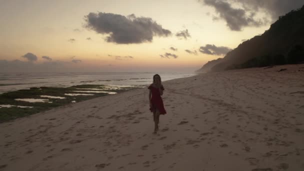 美しい黄金の夕日の間に砂浜を歩く赤いドレスを着た美しい女性の空中ドローンビュー — ストック動画