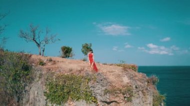 Güneşli yaz gününde deniz kıyısındaki kayalıkta duran kırmızı elbiseli güzel kadının havadan drone görünümü