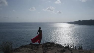 Güneşli yaz günü boyunca deniz kıyısında uçurumda duran kırmızı elbiseli güzel kadının havadan drone geri görünümü