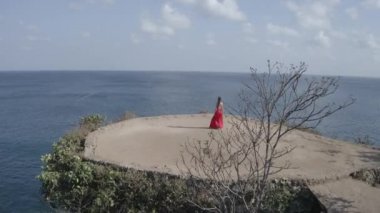 Güneşli yaz gününde deniz kıyısındaki kayalıkta yürüyen kırmızı elbiseli güzel kadının havadan drone görünümü