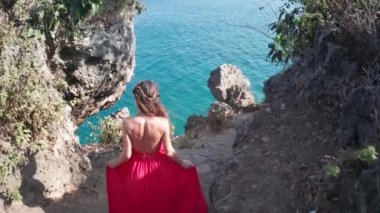 Güneşli yaz gününde deniz kıyısında ki kayalıkta yürüyen kırmızı elbiseli güzel kadının havadan drone geri görünümü